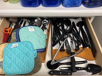 Drawer containing mixed kitchen utensils including serving spoons, knives, tongs, ice cream scoops, and pot holders.