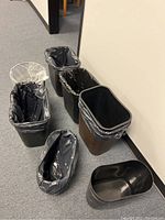 Photo showing a group of assorted plastic trash cans three lined with plastic bags and several unlined, mainly rectangular with black and clear liners visible, in a hallway setting.