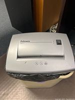 Silver Fellowes PS60-2 paper shredder on beige trash can. Top view shows power switch and paper slot.