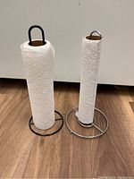 Two metal paper towel holders side by side on a wooden floor against a white wall. One is black with a solid circular base, the other is silver with a 3-spoke base design. Both have paper towels on them.