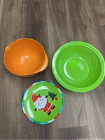 Photo showing three plastic items on a gray wood floor: an orange mixing bowl with a pour spout, a large green bowl, and a colorful plastic plate featuring Santa Claus and Christmas tree design.