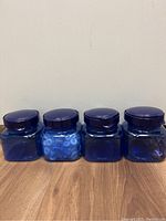 Four cobalt blue square plastic canisters with lids placed side by side on wooden floor against a white wall. One canister contains small blue circular items, others are empty.