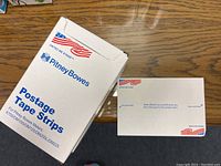 Photo of one open box of Pitney Bowes Postage Tape Strips resting on wooden desk, showing white tape strips with American flag and branding