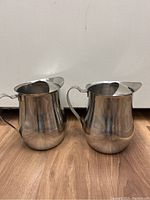 Two polished stainless steel water pitchers placed side by side on wooden flooring in front of a white background, both with handles and pouring spouts visible.