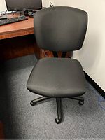 Black task chair with cushioned seat and backrest positioned in an office setting beside a desk with computer equipment