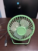 Photo of small green USB desk fan showing front view with metal blades and USB cable.