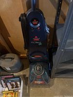 Front view of Bissell ProHeat 2X upright carpet cleaner showing control buttons, brand logos, clear brush head cover and power cord wrapped on the handle.