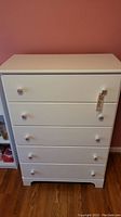 Full frontal view of tall white chest of drawers with five drawers and floral ceramic handles.