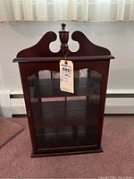 Front view of wooden wall display cabinet with glass door and decorative top finial.