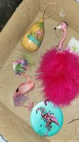Box containing four flamingo themed items including a pink feathered flamingo ornament, small flamingo figurine with flower decoration, flamingo painted gourd, and another pink flamingo ornament.
