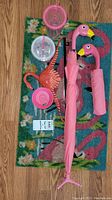 Top view of the lot, showing two flamingo umbrellas with flamingo head handles, pink flamingo hanger, rectangular flamingo door mat, and three plastic cups with lids and straws.