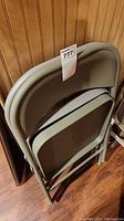 Two gray folding chairs folded and leaning against a wooden wall on a hardwood floor.