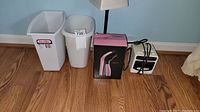 Photo showing 2 white trash cans, pink handheld steamer in box, small alarm clock, and white standing lamp against a blue wall and wood floor.