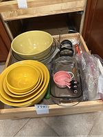 Yellow nested ceramic bowls, clear glass bowls, clear glass rectangular baking tray, pink measuring cups, black and red apple slicer, and small utensils in a kitchen drawer.