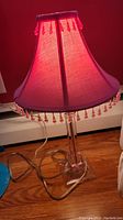 Pink table lamp with fabric shade and beaded fringe, clear glass base with lamp cord and switch visible.