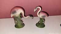 Two ceramic pastel pink flamingo figurines on green bases, one pecking downward, the other standing upright