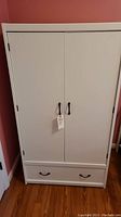 Front view of white wooden wardrobe cabinet with two doors and bottom drawer, showing black handles and overall closed configuration.