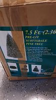 Box close-up showing product label and features: 7.5 Ft (2.30 m) Pre-Lit Scottsdale Pine tree with 1650 natural branch tips, 700 clear pre-strung lights, hinged branches pre-attached, flame retardant, tree stand included.