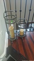 Two pillar candles in metal and glass cylindrical holders on floor near railing.