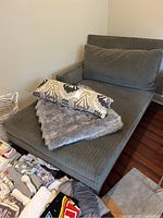 Gray lounge chaise from an upper corner, showing cushions, throw, and surroundings including wooden flooring and adjacent basket.