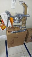 Full view of the artisan Culture brand citrus juicer displayed on the original cardboard box, showing the entire metal structure and lever arm.