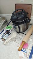 Photo showing Instant Pot 4th Edition multi-cooker, wooden rolling pin, metal baking pan, cookie cutter, and packaging on tiled floor.