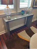 Gray contemporary library table located against a sunlit window with some decorative glass items and a glass wire-frame lamp placed on top for scale and setting.