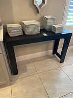 Front angled view of black resin side buffet table with stacks of white rectangular plates on top, table against wall and tile floor visible.
