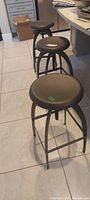 Three black metal stools with round padded seats, shown lined up indoors on a tiled floor.