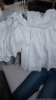 White sleeveless dress with lace trim and a white button-up shirt stacked on couch along with other clothes