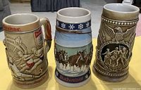 Three beer steins showing detailed painted and embossed designs: Budweiser vintage sports, winter horse wagon, Spirit of 76 historical scene