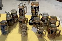 Overview of 12 beer steins in varying sizes and designs, placed on yellow surface showing overall lot contents