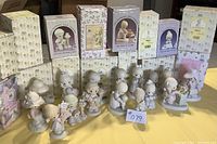 Fifteen Precious Moments figurines arranged in front of their original boxes on a table. Figurines feature children in various poses; one figurine has a broken flower. Boxes show light wear.