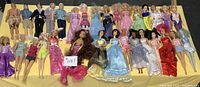 Wide view of 26 female dolls including Barbie and others laid out in rows wearing various dresses and costumes with some hair disheveled.