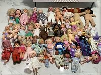 Full view of all dolls laid out on white surface, showing variety of sizes and types mostly vinyl and cloth dolls, some baby dolls, ballerinas and vintage dolls.