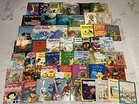 Photo overview showing a large collection of 50 children's soft cover books, arranged in rows with colorful covers visible including animals, holidays, and characters.