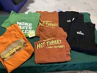 Seven WAWA branded T-shirts in various colors and designs, some with printed slogans and logos laid out on a green surface.