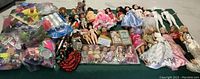 Wide view of multiple dolls and bags of toys, showing variety of dolls with some dressed and others loose, assorted toy accessories included.