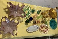 Photo showing two large unique-shaped Murano Art Deco style bowls, several small glasses in green, blue, pink, amber, swirl patterned candy dishes, amber candle holders, blue glass plate, long white dish, and a tall amber wine bottle with shot glasses on a yellow tablecloth.