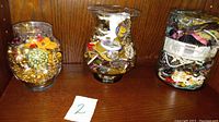 Three large glass jars filled with various costume jewelry pieces such as necklaces, bracelets, rings, and earrings.