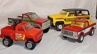 Four vintage metal toy trucks arranged together showing red Tonka truck, red and silver pickup, red and green Nylint Wilderness Camper, yellow and black Turbo Blazer