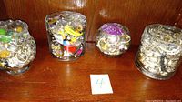 Four small glass jars filled with assorted vintage costume jewelry displayed on wooden surface.