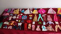 Photo showing a spread of over 75 vintage Barbie clothes on a dark red surface, including dresses, jackets, pants, shirts, skirts, and accessories with bright and pastel colors.