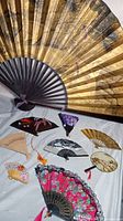 Nine Asian fans of various sizes and materials displayed on a white cloth background