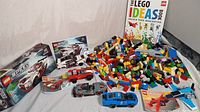 Photo of four assembled LEGO cars, a small LEGO plane, a large pile of LEGO bricks, LEGO Ideas book, and LEGO Speed Champions Nissan GT-R NISMO set box with instruction booklet.