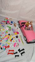 Photo showing pink Barbie Rolls Royce convertible with two dolls inside, surrounded by various colorful doll accessories including shoes, hats, purses, sunglasses, and toy dogs.