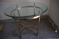 Photo of a brass base with oval clear glass top side table on carpeted floor near wall showing brass ring and finials