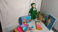 Wide view of the hanging clown doll in green sequin outfit on a wooden seat, two vintage doll books standing against the wall, paper doll set in box, and the Kenner playset with accessories laid out on the floor.