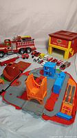 Overview of the playset featuring the Fisher Price fire station, multiple toy vehicles including Nylint fire truck, and several small scale toy cars grouped on the gray base with orange and blue plastic details.