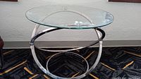 Front view of the round glass side table showcasing the polished chrome geometric base and clear glass top on a black and gold patterned carpet.